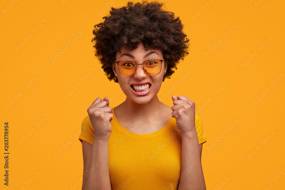 Portrait of annoyed female teacher with Afro hairstyle, clenches teeth and fists angrily, being irritated with naughty pupils, wears trendy clothes, isolated over yellow background. Negative emotions