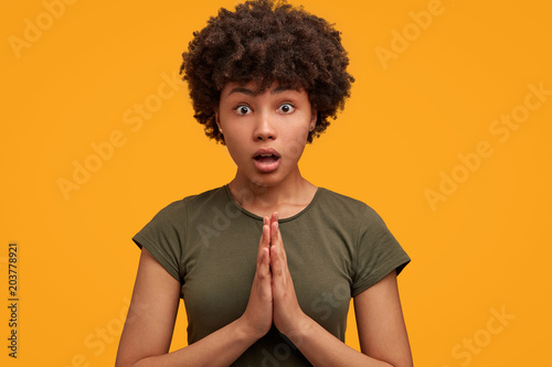 Shocked beautiful African American female feels worried and excited, presses palms together, being terrified with something, poses against yellow background. People and surprisement concept.