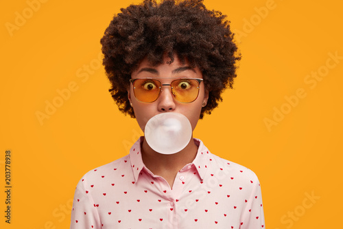 Playful funny African American woman blows chewing gum, spends spare time with friends, wears shirt and sunglasses, isolated over yellow background. Shocked female inflates bubble gum in studio photo