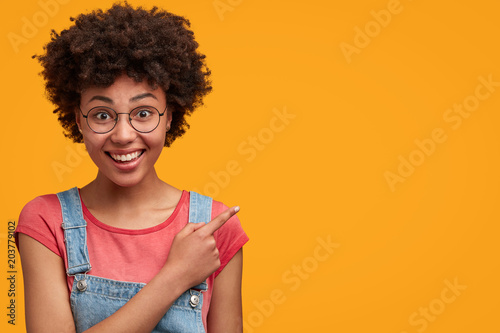 Happy clever African college student with broad smile, indicates at blank copy space for your promotional text, has delighted expression, isolated over yellow background. Advertisement concept