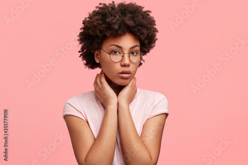 Headshot of pleasant looking mixed race young female has dark curly hair, looks confidently at camera, has healthy dark skin, wears casual t shirt and glasses, poses against pink background.