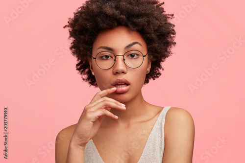 Close up shot of thoughtful lovely young African American female with dark healthy skin looks pensively directly at camera, thinks about something, poses over pink studio background. Beauty, ethnicity