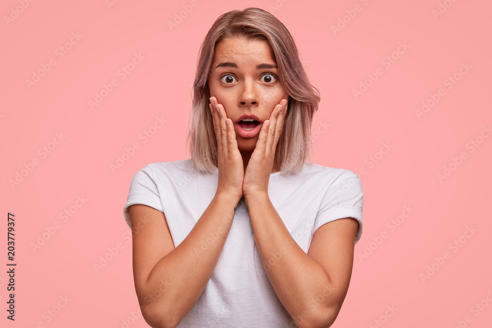 Studio shot of shocked female looks with unexpected expression, finds out about bad results on exam, keeps jaw dropped, wears casual white t shirt, poses in pink studio alone. Astonished woman Studio shot of shocked female looks with unexpected expression, finds out about bad results on exam, keeps jaw dropped, wears casual white t shirt, poses in pink studio alone. Astonished woman