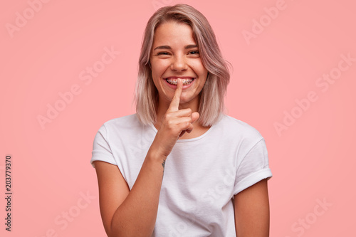 Pretty female with broad smile, demonstrates teeth with brackets, shows silence sign, wears casual white t shirt, isolated over pink background. Glad European woman has bobbed haircut, tells secret
