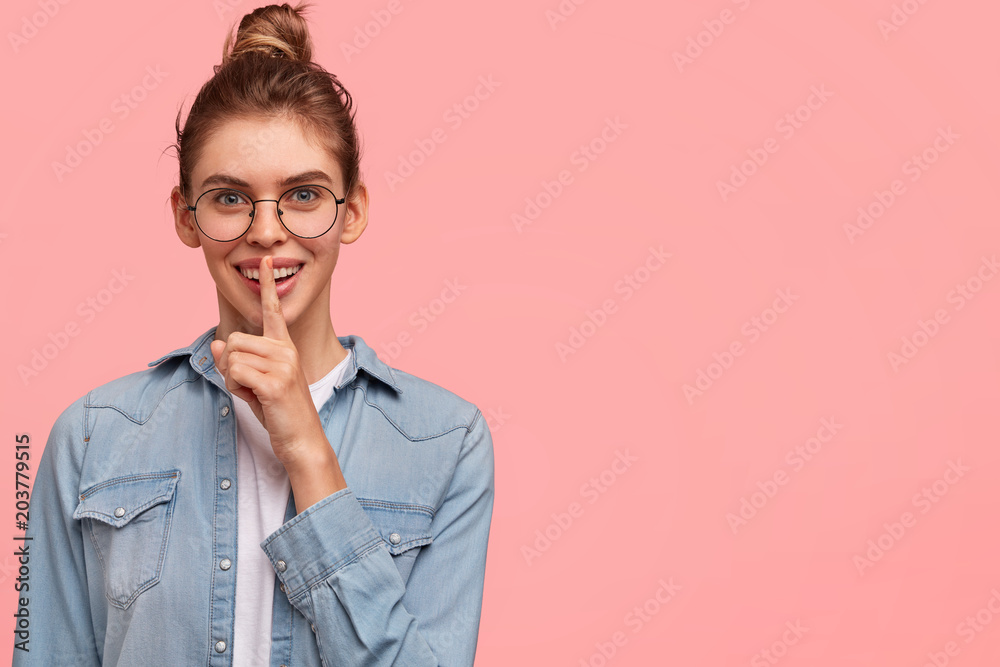 Pleased attractive young woman with happy smile, shows hush gesture, asks to keep confidential information in secret, poses against pink studio background with copy space for your advertisement Pleased attractive young woman with happy smile, shows hush gesture, asks to keep confidential information in secret, poses against pink studio background with copy space for your advertisement
