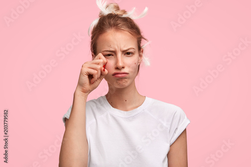 Sleepy female rubs eyes, didn`t have enough sleep, worked for all night, has feather on head, wears casual t shirt, poses against pink background. Upset young Caucasian woman slumbers in bedroom photo