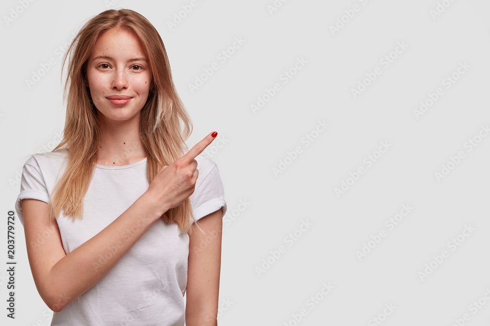 Portrait of attractive female with pleasant appearance, advertises something at blank white copy space on white background. Horizontal shot of lovely young woman indicates with index finger. Portrait of attractive female with pleasant appearance, advertises something at blank white copy space on white background. Horizontal shot of lovely young woman indicates with index finger.