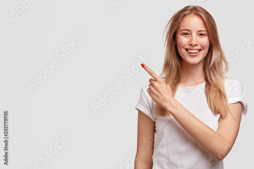 Positive Caucasian female in casual t shirt, indicates at blank copy space fo...