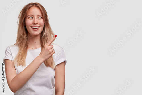 Image of content beautiful female with pleasant smile, shows with index finger at blank copy space for your advertising content, wears casual white t shirt. People, happiness and advertisement