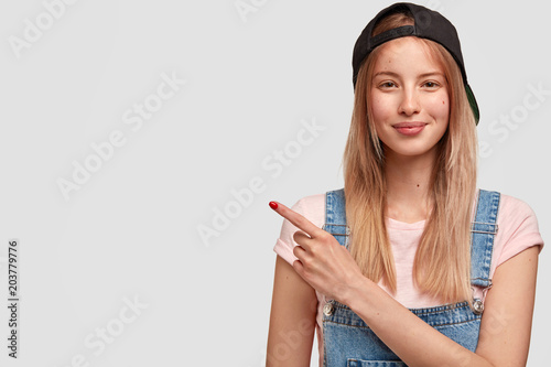 Happy female youngster in trendy black cap and denim overalls, has delighted expression, lovely appearance, indicates at blank copy space for your advertisement or promotional text. Look there