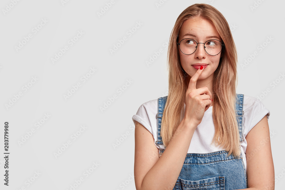 Pensive gorgeous good looking female model looks thoughtfully upwards, wears spectacles and denim overalls, poses against white background with copy blank space for your advertising content. Pensive gorgeous good looking female model looks thoughtfully upwards, wears spectacles and denim overalls, poses against white background with copy blank space for your advertising content.