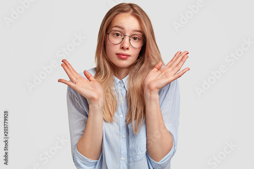 Uncertain puzzled young cute female pupil shrugs shoulders, feels unsure while answers on final exam, looks hesitant and says: Who cares, I don`t know. People, reaction and body language concept