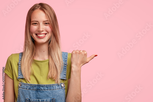 Photo of delighted smiling female student being in good mood  rejoices successfully passed exam  points with thumb at blank copy space for your advertisement. People  positive emotions concept