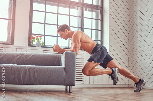 Handsome shirtless muscular male does exercise leaning on a sofa at home.