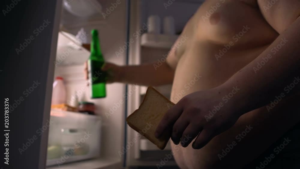 Addicted to fast food fat man eating sandwich and drinking beer near fridge Addicted to fast food fat man eating sandwich and drinking beer near fridge