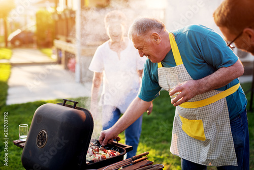 Elderly man is carefully inspecting should he take meat off the grill while his wife is standing beside him and laughing.