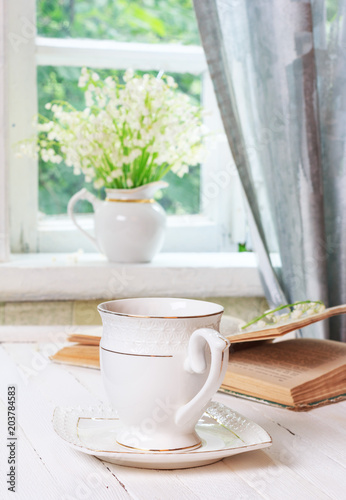 a cup of tea or coffee and a book on a white wooden antique retro table and a bouquet of lily-of-the-valley flowers on a window sill near an open window in a country house in the spring morning