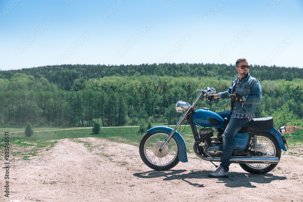 Portrait young guy with sunglasses and Jeans jacket and pants posing sitting on his vintage retro motorbike. biker wearing jeans, fashion men. old timer age concept, 1960s style, outdoor dirt road Portrait young guy with sunglasses and Jeans jacket and pants posing sitting on his vintage retro motorbike. biker wearing jeans, fashion men. old timer age concept, 1960s style, outdoor dirt road