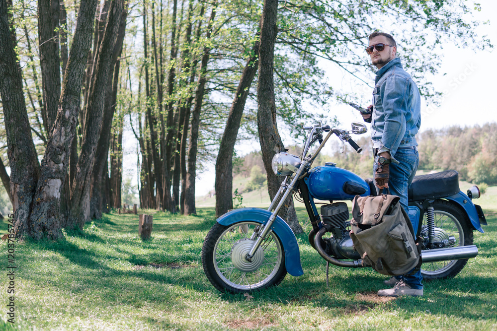 Portrait young guy with sunglasses and Jeans jacket and pants posing sitting on his vintage retro motorbike. biker wearing jeans, fashion men. old timer age concept, 1960s style, outdoor dirt road
