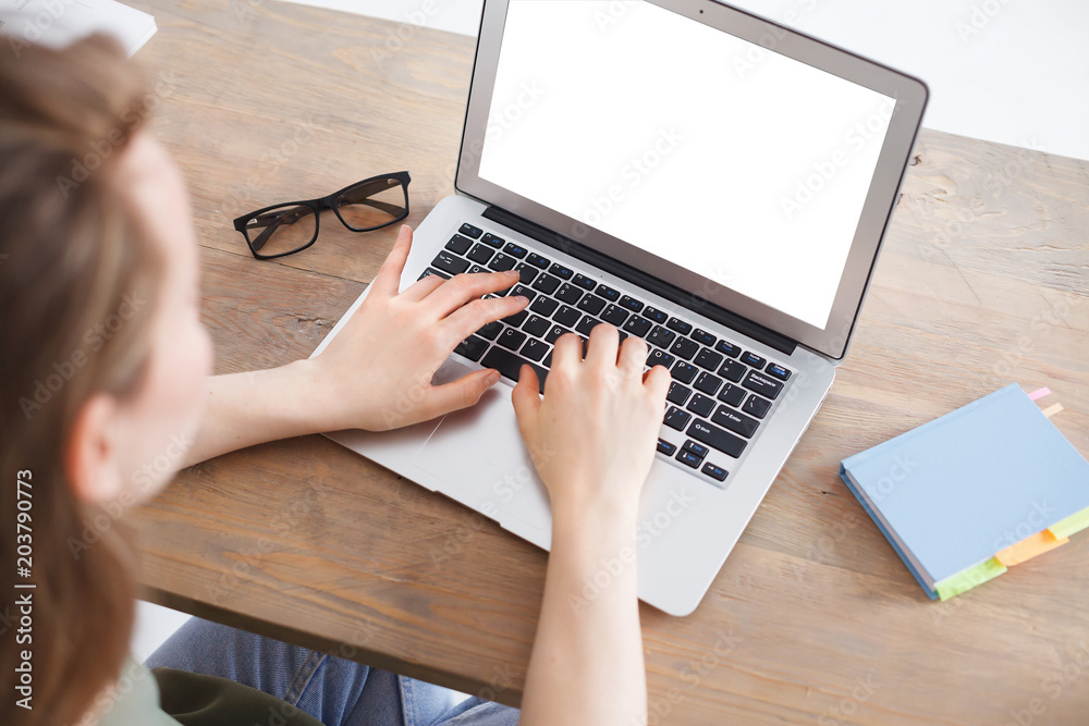 Fototapeta premium Top view of female hands typing on computer keyboard.Woman working on laptop sitting at work table. Glasses and Notes lying on wooden table. Laptop computer with blank screen for your promotional text