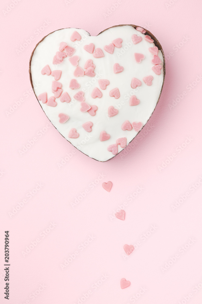 White heart shaped muffin decorated with little pink sugar hearts on pink background. Vertical image.