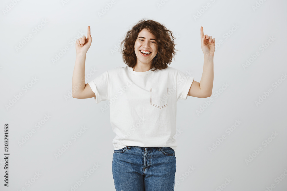 Make daily routine bright with this copy space. Portrait of joyful ordinary european woman with short curly hair, raising index fingers and pointing up, smiling and laughing from happiness Make daily routine bright with this copy space. Portrait of joyful ordinary european woman with short curly hair, raising index fingers and pointing up, smiling and laughing from happiness