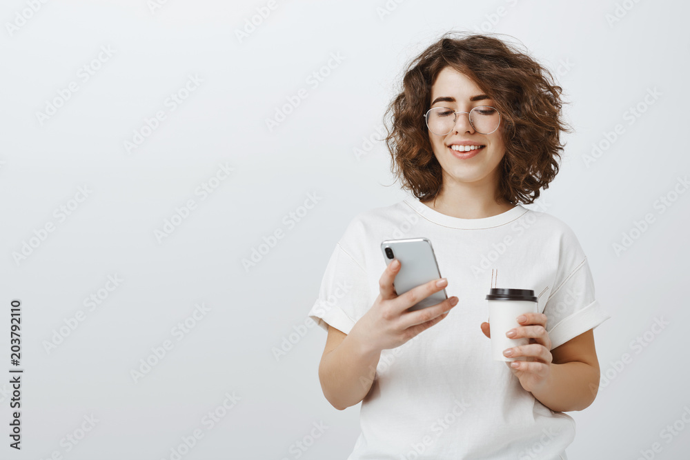 Busy cute businesswoman checking schedule. Portrait of satisfied successful designer in glasses, holding cup of coffee and holding smartphone, smiling while looking at screen happily Busy cute businesswoman checking schedule. Portrait of satisfied successful designer in glasses, holding cup of coffee and holding smartphone, smiling while looking at screen happily