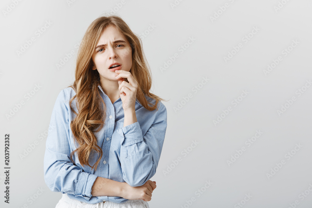 Trendy girl having doubts about new dress. Studio shot of disbelieving good-looking female student in blue-collar shirt, touching lip and staring with concerned expression, thinking over gray wall Trendy girl having doubts about new dress. Studio shot of disbelieving good-looking female student in blue-collar shirt, touching lip and staring with concerned expression, thinking over gray wall