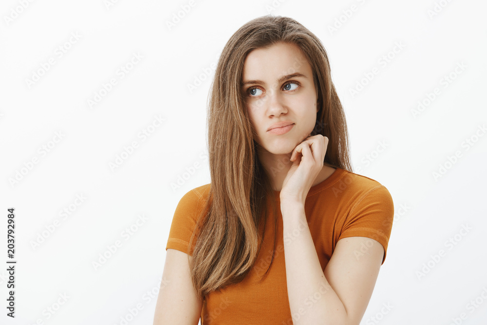 Portrait of troubled good-looking creative woman with fair hair in mustard t-shirt, rubbing chin and looking left with displeased worried expression, thinking or trying to solve problem over gray wall Portrait of troubled good-looking creative woman with fair hair in mustard t-shirt, rubbing chin and looking left with displeased worried expression, thinking or trying to solve problem over gray wall