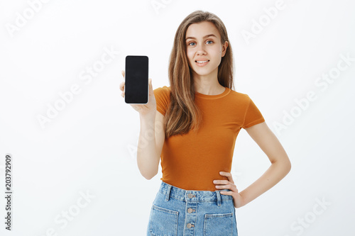Show me better device. Good-looking confident woman with blond hair, holding hand on hip and showing smartphone, advertising or discussing with friends gift from parent for good marks in university