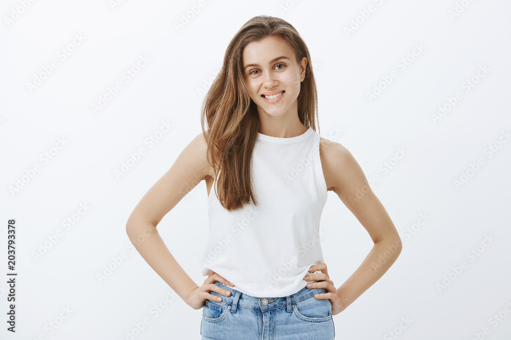 Obraz premium Confident attractive feminine girlfriend with fair hair in stylish tank-top, holding hands on hips and smiling joyfully at camera, satisfied with success, having great time while hanging out in pub