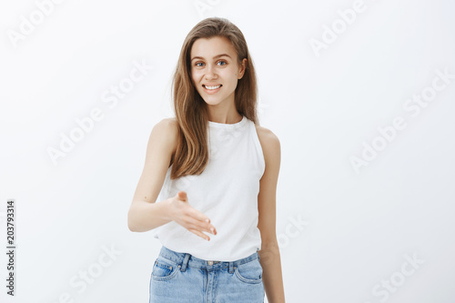 Indoor shot of pleasant good-looking feminine girl with fair hair, pulling palm towards camera to give handshake and smiling cheerfully, greeting business partner and standing confident over gray wall © Cookie Studio