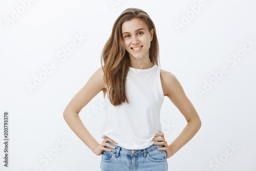 Confident attractive feminine girlfriend with fair hair in stylish tank-top, holding hands on hips and smiling joyfully at camera, satisfied with success, having great time while hanging out in pub