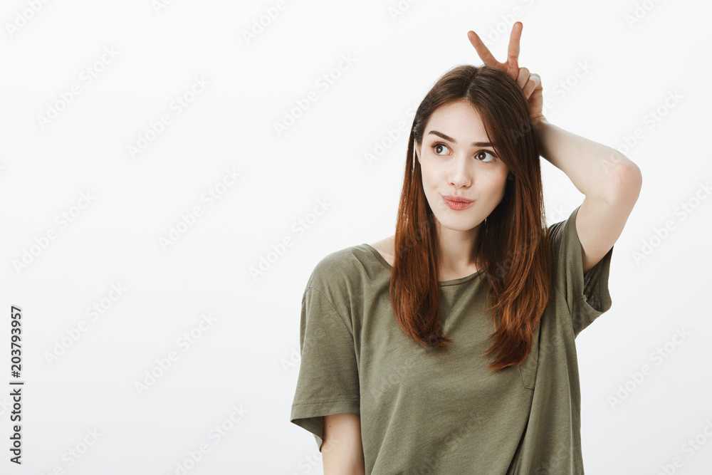 Girl mimicking easter bunny, being in good dreamy mood. Attractive female student in green t-shirt, looking aside with focused cute expression, holding v sign behind head as if showing animal ears