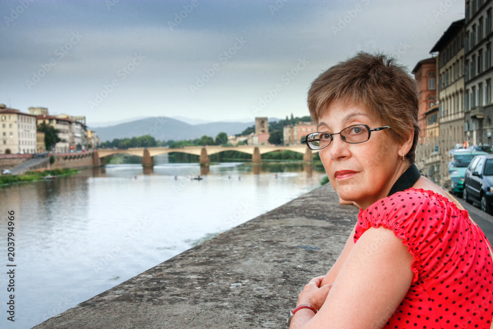 Mature tourist woman travelling in Italy stays against bridge over Arno river in Florence.