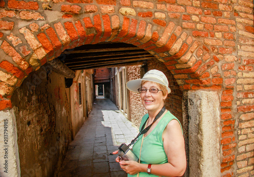 Happy mature tourist woman travelling in Italy stays against narrow street and arch in Venice.