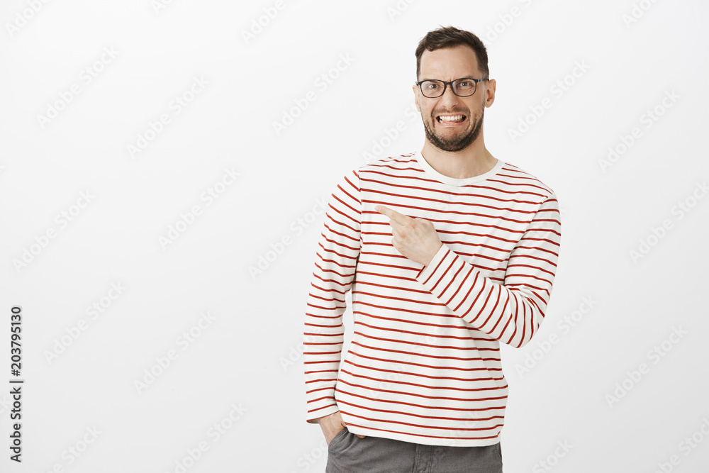 Indoor shot of disgusted and displeased funny guy in glasses, grimacing from awful smell, pointing at upper left corner while expressing negative opinion and dislike over gray background Indoor shot of disgusted and displeased funny guy in glasses, grimacing from awful smell, pointing at upper left corner while expressing negative opinion and dislike over gray background