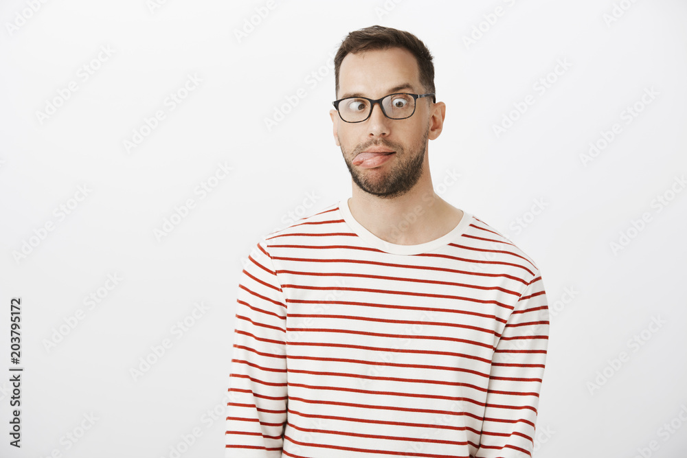 Portrait of playful funny guy with bristle in black eyewear, sticking out tongue and looking at nose, making faces and being childish over gray background, being bored and emotive Portrait of playful funny guy with bristle in black eyewear, sticking out tongue and looking at nose, making faces and being childish over gray background, being bored and emotive