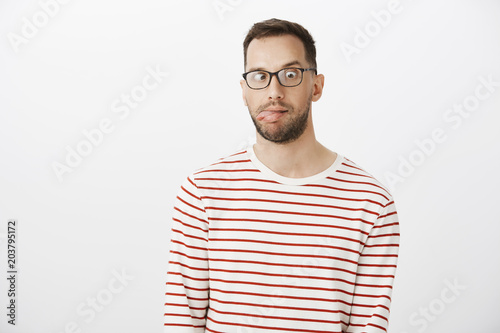 Portrait of playful funny guy with bristle in black eyewear, sticking out tongue and looking at nose, making faces and being childish over gray background, being bored and emotive © Cookie Studio