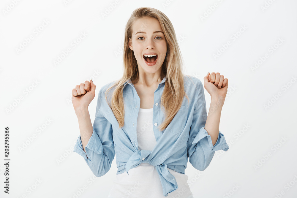 Positive triumphing charming woman with blond hair, raising hands and clenching fists from pleased excitement, smiling broadly, celebrating success or victory, being pleased with good result Positive triumphing charming woman with blond hair, raising hands and clenching fists from pleased excitement, smiling broadly, celebrating success or victory, being pleased with good result