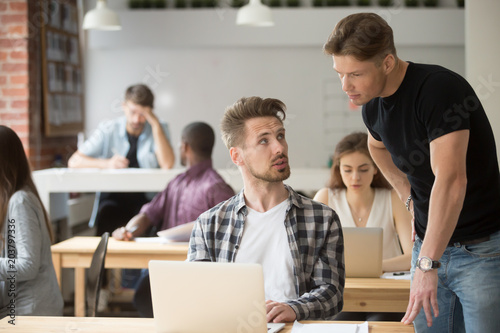 Male intern asking for help from experienced supervisor showing problem on laptop in coworking office space. Two male colleagues consulting and negotiating on work issues. Concept of help, cooperation