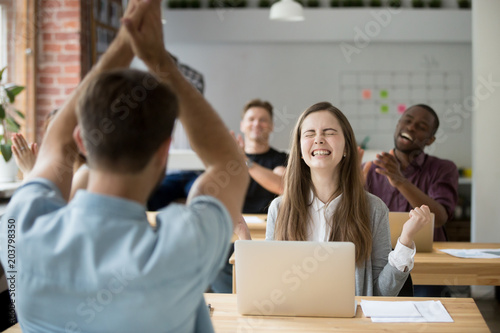Excited female worker gesturing Yes sign, happy with good business news on stock market, online win, startup success with colleagues applauding and congratulating. Concept of rewarding, achievement