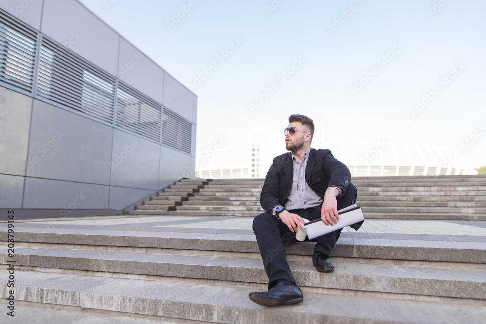 Portrait of a business man sitting on the street with coffee and documents in his hands and looking away. Businessman sitting on a staircase against a background of a city landscape. Portrait of a business man sitting on the street with coffee and documents in his hands and looking away. Businessman sitting on a staircase against a background of a city landscape.