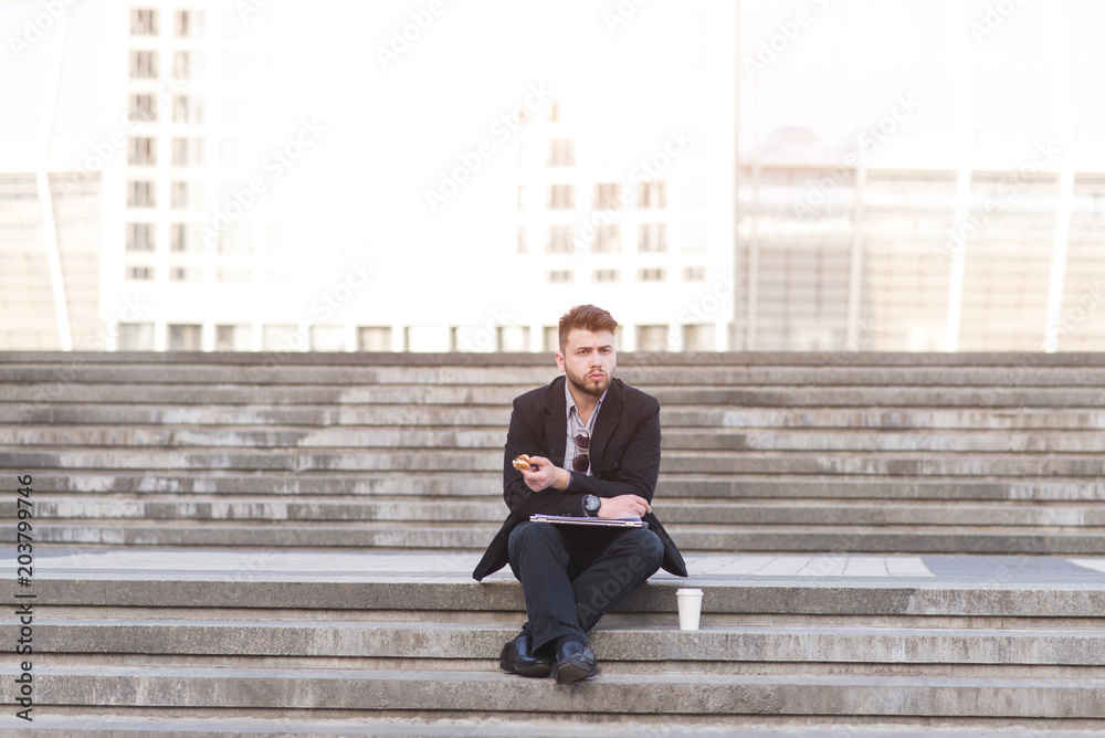 Busy man is sitting on the street with a snack and a cup of coffee in the background of the city landscape. Businessman sitting on the stairs and eating. Busy man is sitting on the street with a snack and a cup of coffee in the background of the city landscape. Businessman sitting on the stairs and eating.