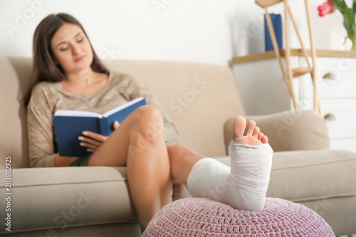 Fotografia Young woman with broken leg in cast reading book while sitting on sofa at home