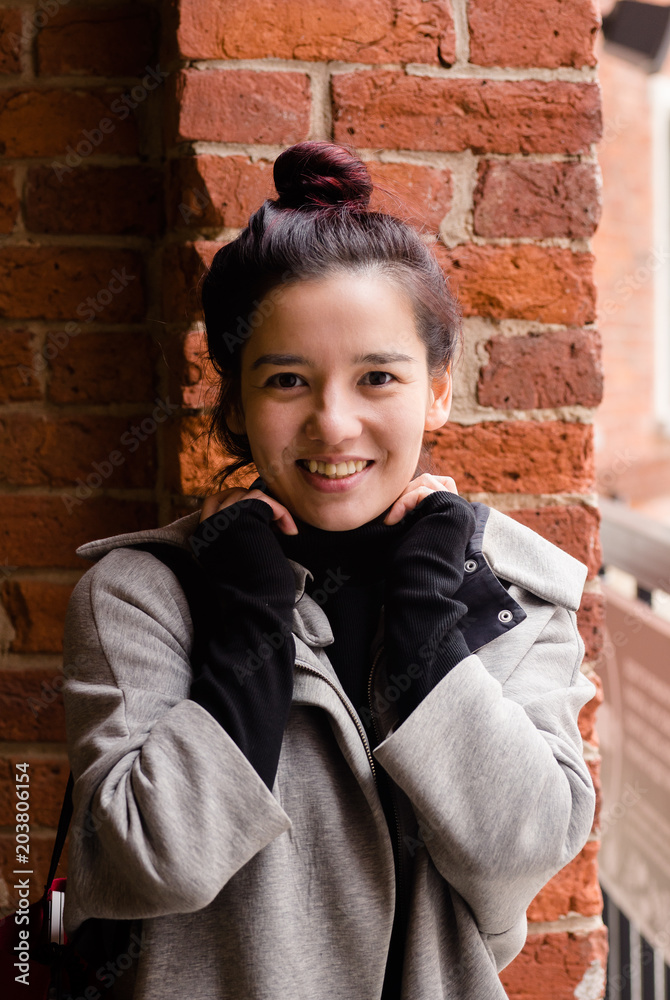 Image of young beautiful female model of mixed race standing next to the brick wall dressed in gray coat with red textile bag. Image of young beautiful female model of mixed race standing next to the brick wall dressed in gray coat with red textile bag.
