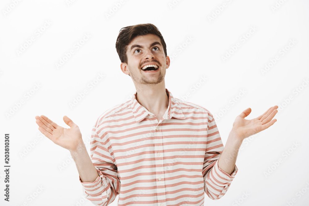 God what next. Portrait of sarcastic laughing boyfriend in stripes shirt, raising hands up and looking at sky with smile, seeing something hilarious and interesting, standing against gray background God what next. Portrait of sarcastic laughing boyfriend in stripes shirt, raising hands up and looking at sky with smile, seeing something hilarious and interesting, standing against gray background