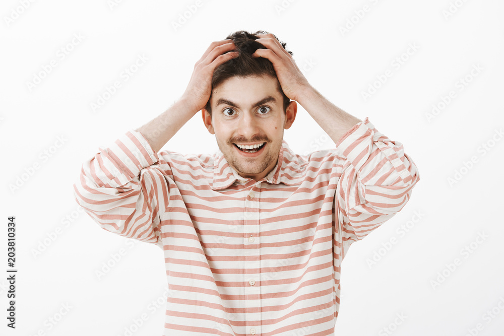 Indoor portrait of fascinated lucky male model with moustache, holding hands on hair and smiling happily, doubting his own luck, standing against gray background overwhelmed with great emotions Indoor portrait of fascinated lucky male model with moustache, holding hands on hair and smiling happily, doubting his own luck, standing against gray background overwhelmed with great emotions