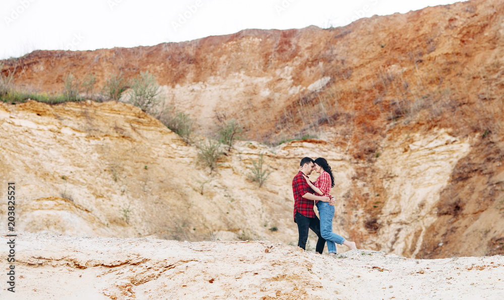 Lovely couple in red plaid shirts standing and embrace in sand canyon or quarry with mountain on the background. Outdoor