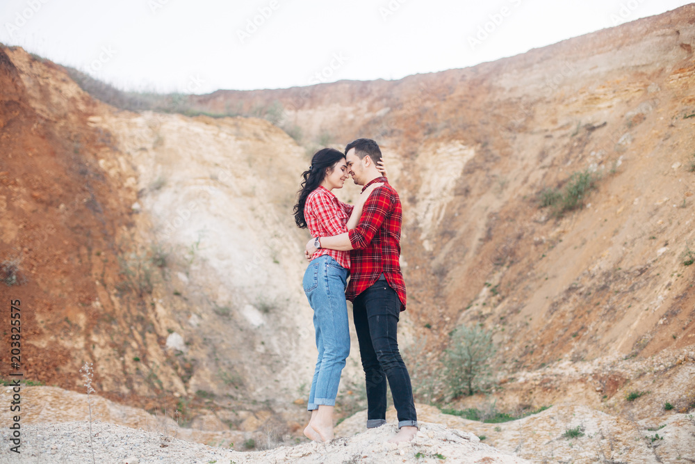 Lovely couple in red plaid shirts standing and embrace in sand canyon or quarry with mountain on the background. Outdoor